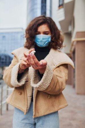 Portrait of young girl with curly hair in protective mask that using antiseptic outdoors near business building at quarantine time. Conception of coronavirusの写真素材