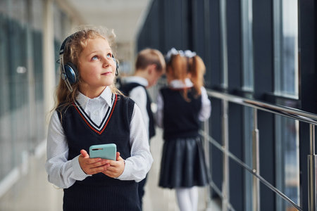 Little girl with phone and headphones standing if front of school kids in uniform that together in corridor. Conception of educationの写真素材