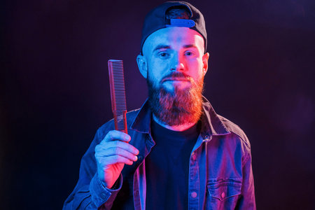 Young bearded barber in cap standing in the studio with neon lightingの写真素材