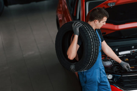 Worker in black and blue uniform holds car wheel and walks with in near red automobileの写真素材