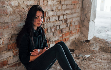Young beaten woman in black clothes sitting on the floor of abandoned buildingの写真素材