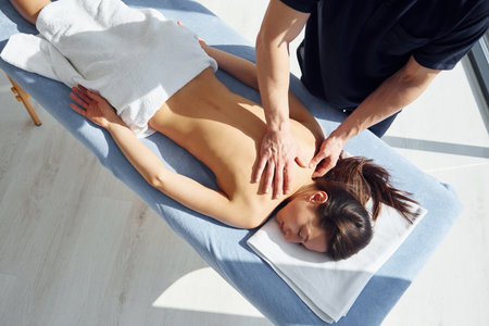 Room illuminated by sun beams. Young woman is lying down when man doing massage of her body at spaの写真素材