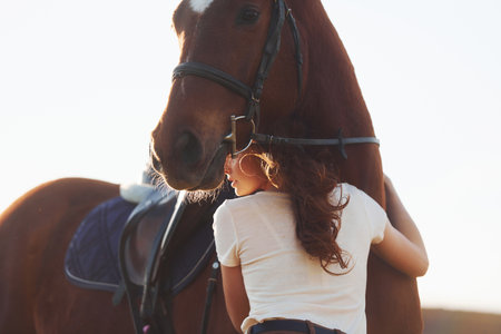 Beautiful sunshine. Young woman standing with her horse in agriculture field at daytimeの写真素材