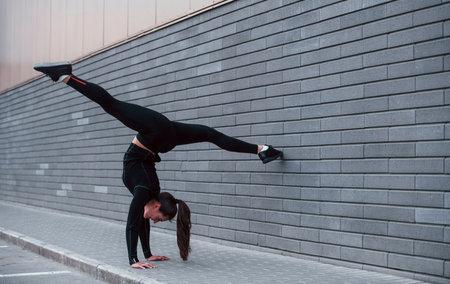 Young sportive girl in black sportswear doing hard handstand exercises outdoors near gray wallの写真素材