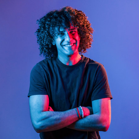 Posing for a camera. Young beautiful man with curly hair is indoors in the studio with neon lightingの写真素材
