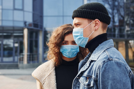 Couple in protective masks have a walk outdoors in the city near business building at quarantine time. Conception of coronavirusの写真素材