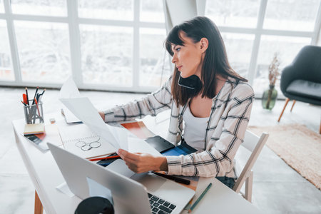 Sits by table. Young female freelance worker is indoors in home at daytimeの写真素材