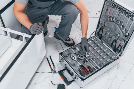 Young professional plumber in grey uniform using equipment on the kitchen and preparing for the workの写真素材