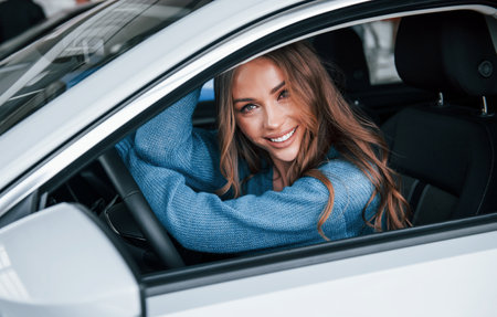 Positive woman in blue shirt sits inside of new brand new car. In auto salon or airportの写真素材