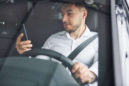 Successful man. Young guy in white shirt is sitting inside of a modern new automobile