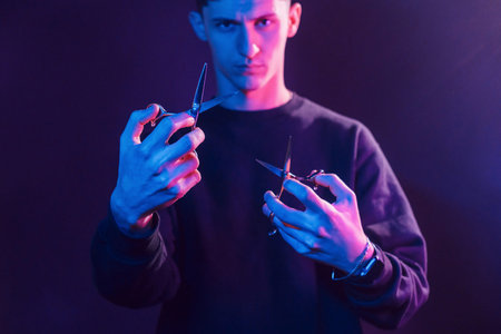 Holds scissors. Young barber with work equipment standing in the studio with neon lighting