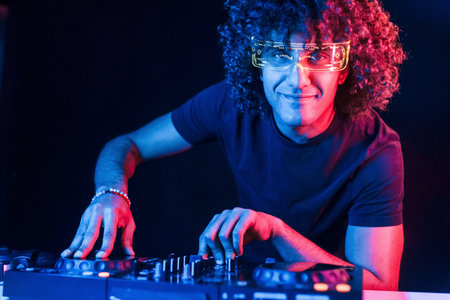 Man with curly hair using DJ equipment and standing in the dark neon lighted roomの写真素材