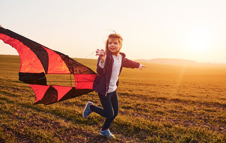 Happy little girl running with kite in hands on the beautiful field at sunrishe timeの写真素材