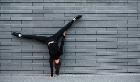 Young sportive girl in black sportswear doing hard handstand exercises outdoors near gray wallの写真素材