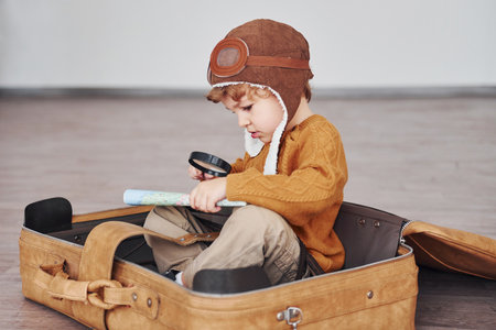 Little boys in retro pilot costume have fun and sitting in suitcase indoors at daytimeの写真素材