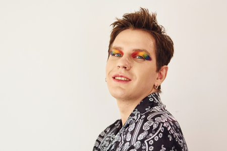 Young gay man is standing in the studio and posing for a camera. Multicolored make up on face fingersの写真素材