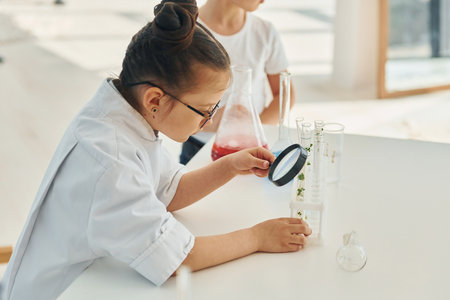 Holds magnifying glass. Little girl in coat playing a scientist in lab by using equipmentの写真素材