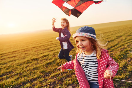Two little girls friends have fun together with kite and toy car on the field at sunny daytimeの写真素材