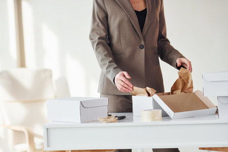 Packaging orders into the white boxes. Young woman in formal clothes is indoors. Conception of styleの写真素材