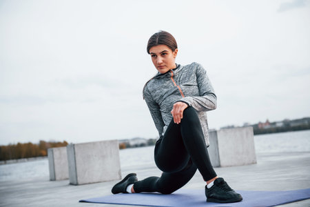 Young sportive girl doing yoga exercises on fitness mat outdoors near lake at daytime