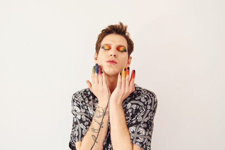 Young gay man is standing in the studio and posing for a camera. Multicolored make up on face fingersの写真素材