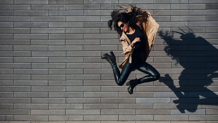 Beautiful brunette with curly hair and in black clothes jumping and having fun outdoors near wallの写真素材