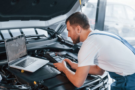 Uses laptop. Young man in white shirt and blue uniform repairs automobileの写真素材