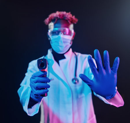 Smart doctor in protective uniform holding infrared thermometer. Futuristic neon lighting. Young african american man in the studioの写真素材