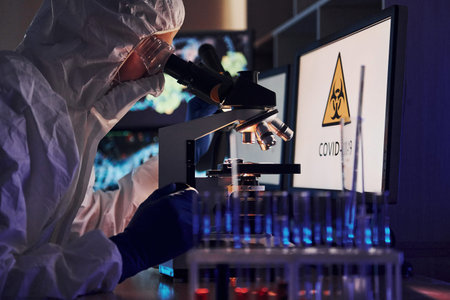 Monitors with information on the table. Scientist in white protective uniform works with coronavirus...