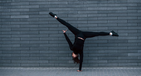 Young sportive girl in black sportswear doing hard handstand exercises outdoors near gray wallの写真素材