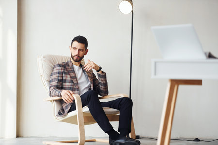 Sits by table in office. Young stylish businessman in suit indoors. Conception of successの写真素材