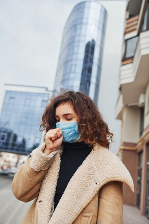 Portrait of young girl with curly hair in protective mask that have a cough outdoors near business building at quarantine time. Conception of coronavirusの写真素材