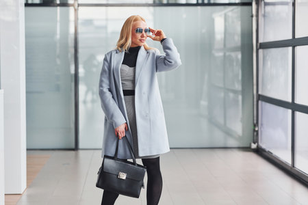 Beautiful blonde in elegant clothes with black handbag standing indoors against grey backgroundの写真素材