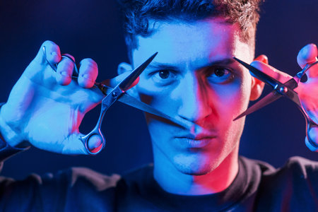 Holds scissors. Young barber with work equipment standing in the studio with neon lightingの写真素材