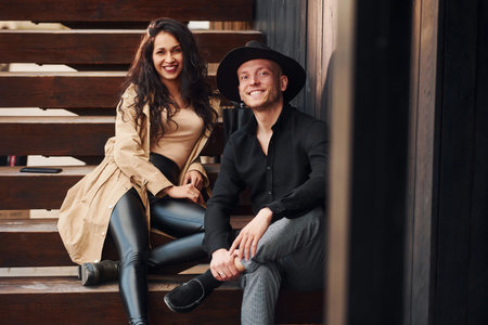 Cheerful brunette in black leggings sitting on wooden stairs with her man in hatの写真素材