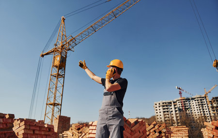 Talking by phone. Construction worker in uniform and safety equipment have job on buildingの写真素材