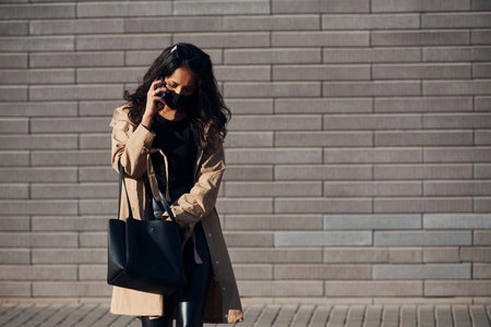 In protective mask. Beautiful brunette with curly hair and in black clothes talking by phone outdoors near wallの写真素材