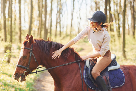 Young woman in protective hat with her horse in the forest at sunny daytimeの写真素材