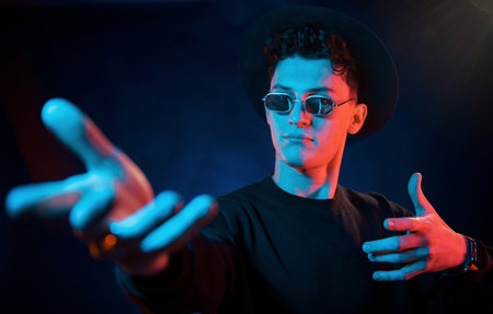 In hat and glasses. Neon lighting. Young european man is in the dark studioの写真素材
