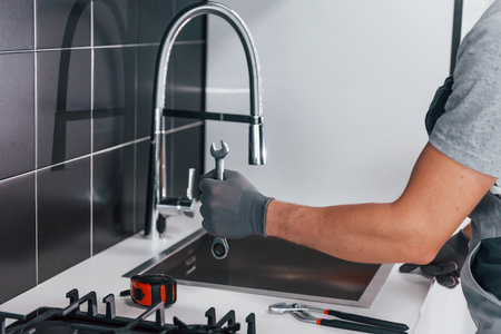 Close up view of young professional plumber in grey uniform holding wrench in hand on the kitchenの写真素材