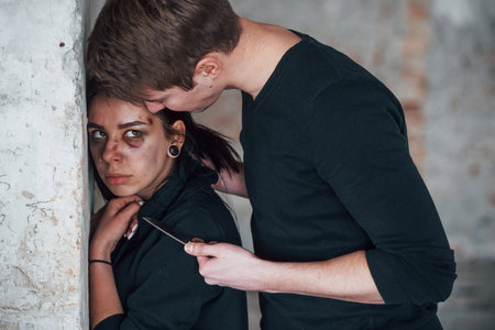 Kidnapped woman standing near wall and threatened by man with knife in hands in abandoned buildingの写真素材