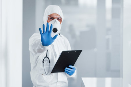 Shows stop gesture by hand. Male doctor scientist in lab coat, defensive eyewear and mask holding notepad in handsの写真素材