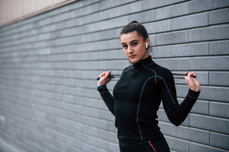 Young sportive girl in black sportswear standing with jump rope in hands outdoors near gray wallの写真素材