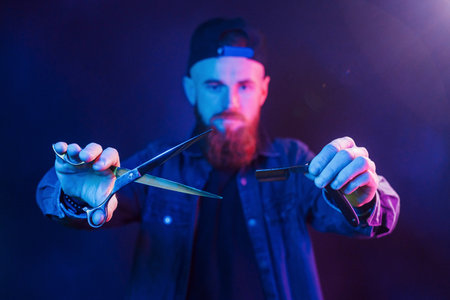 Young bearded barber in cap standing in the studio with neon lightingの写真素材
