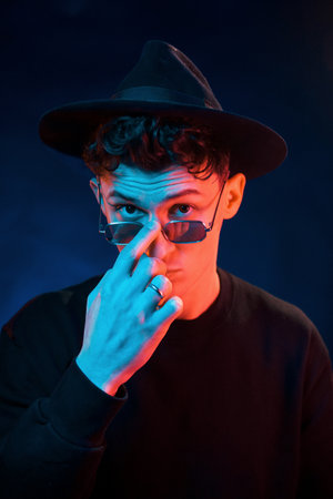 In hat and glasses. Neon lighting. Young european man is in the dark studioの写真素材