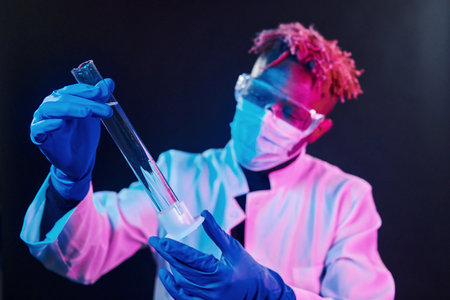 Smart scientist in protective uniform holding test tube. Coronavirus conception. Futuristic neon lighting. Young african american man in the studioの写真素材