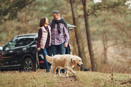 Happy mature couple have a walk with their dog in autumn or spring forest near modern carの写真素材