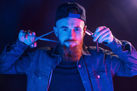 Young bearded barber in cap standing in the studio with neon lightingの写真素材
