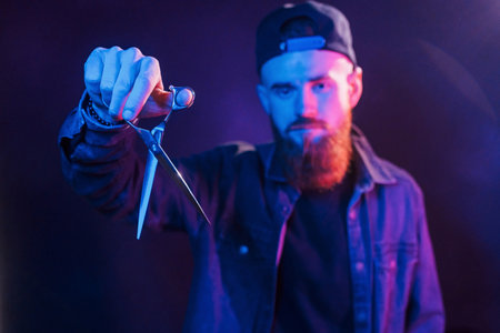 With scissors. Young bearded barber in cap standing in the studio with neon lightingの写真素材