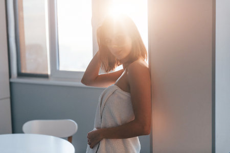 Standing at sunrise time in white towel. Young woman is indoors in room of modern house at daytimeの写真素材